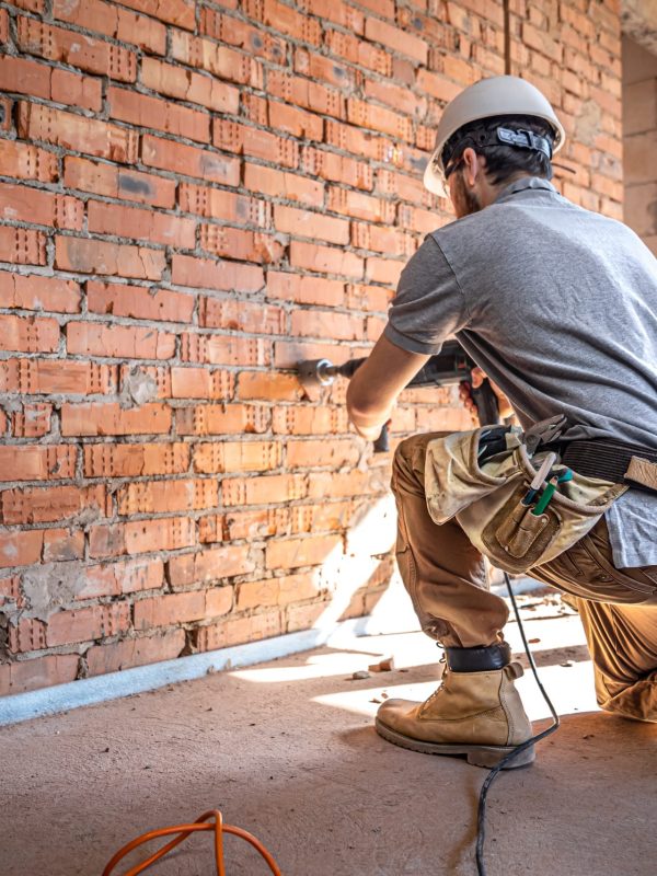 A builder works with a drill at a construction site. Handyman at a construction site in the process of drilling a wall with a perforator.